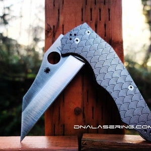 Puede incluir: Un cuchillo de bolsillo plateado con un mango de patrón blanco y negro. El mango tiene un diseño geométrico de círculos superpuestos. El cuchillo está abierto y la hoja es visible. El cuchillo está apoyado sobre una superficie de madera.