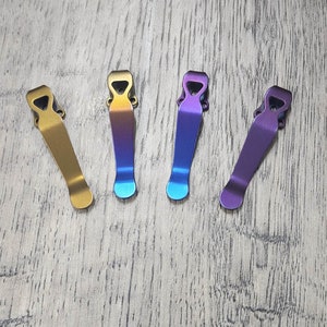 May include: Four metal pocket clips in gold, blue-purple, blue-green, and purple hues. Each clip has a curved design with a small opening at the top and a rounded bottom. The clips are arranged on a light gray wooden surface.
