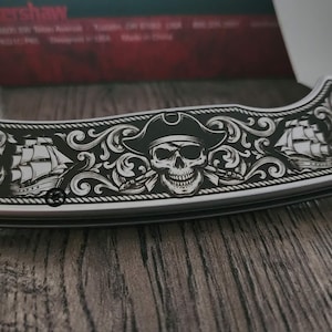 Puede incluir: Una navaja plateada con un dise&ntilde;o detallado de tem&aacute;tica pirata. El mango presenta una calavera con sombrero, barcos, anclas y una br&uacute;jula. La hoja es parcialmente visible y la navaja descansa sobre una superficie de madera.