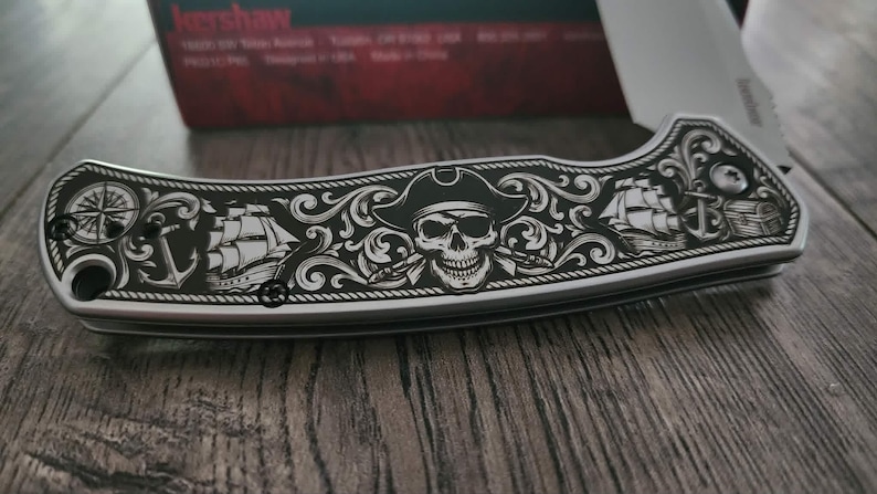 Puede incluir: Una navaja plateada con un dise&ntilde;o detallado de tem&aacute;tica pirata. El mango presenta una calavera con sombrero, barcos, anclas y una br&uacute;jula. La hoja es parcialmente visible y la navaja descansa sobre una superficie de madera.
