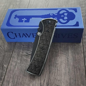 May include: A black folding knife with a patterned handle rests on a wooden surface, next to a blue box. The box features a key design and the text "CHAVE KNIVES". The knife has a silver blade and a textured handle.