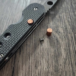 May include: A black folding knife with a copper colored pin and a small screw on a wooden surface.