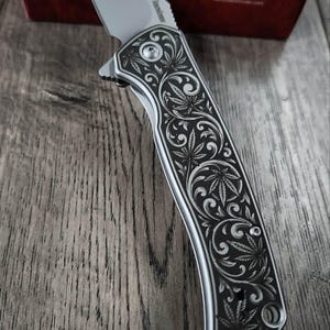 May include: A silver folding knife with a leaf design on the handle. The blade is open, and the handle is detailed with an engraved pattern. A red and black box with the brand name "Kershaw" is in the background.