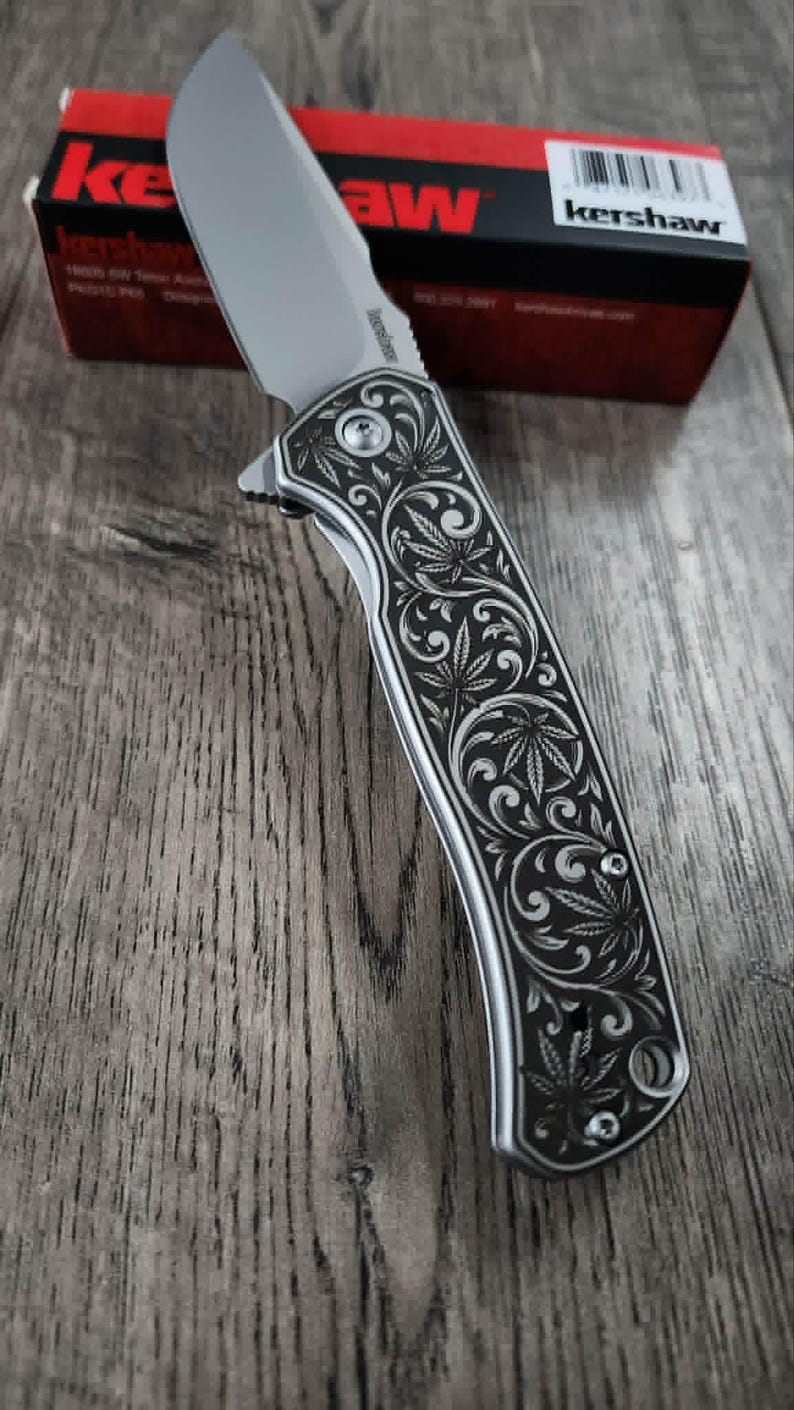 May include: A silver folding knife with a leaf design on the handle. The blade is open, and the handle is detailed with an engraved pattern. A red and black box with the brand name "Kershaw" is in the background.