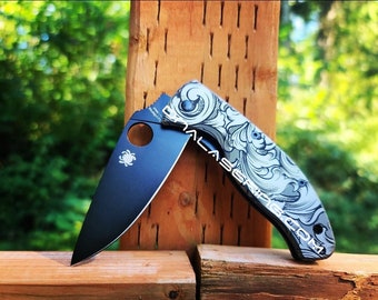 May include: A black folding knife with a silver patterned handle. The knife is open and the blade is visible. The knife is resting on a wooden surface.