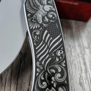 May include: A silver folding knife with a detailed engraving of an eagle and the American flag. The handle features ornate scrollwork and is secured with small screws. The blade is partially visible, with the brand name "Kershaw" etched on it.