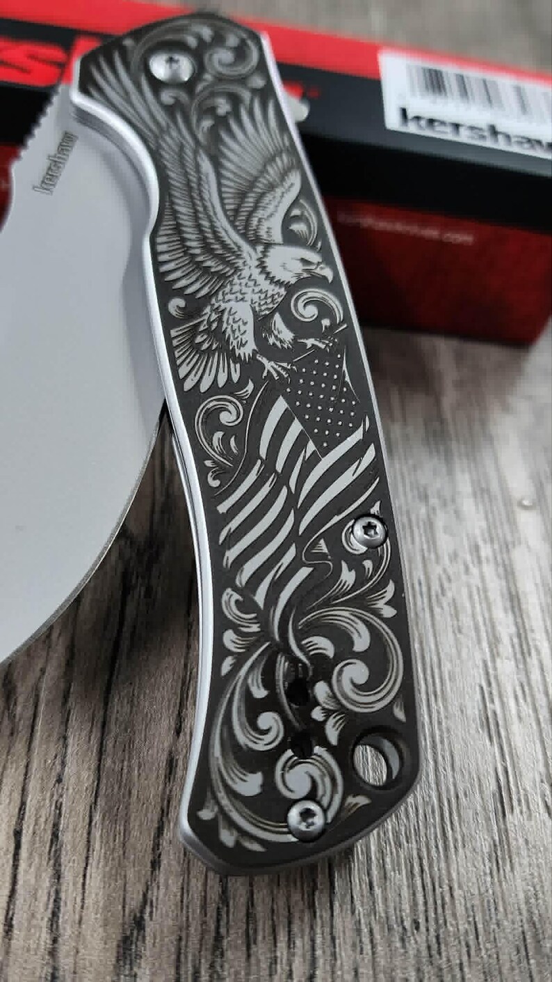 May include: A silver folding knife with a detailed engraving of an eagle and the American flag. The handle features ornate scrollwork and is secured with small screws. The blade is partially visible, with the brand name "Kershaw" etched on it.
