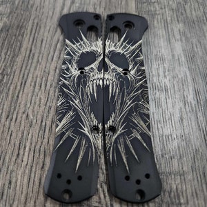 May include: Two black knife scales with a detailed, engraved skull design. The skull features sharp teeth and intricate patterns, with a spiky crown-like element. The scales have holes for screws and a smooth, matte finish.