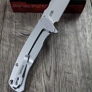 May include: A silver Kershaw folding knife with a partially serrated blade, on a wooden surface. The knife has a pocket clip and the brand name is visible. A red and black box with the Kershaw logo is in the background.