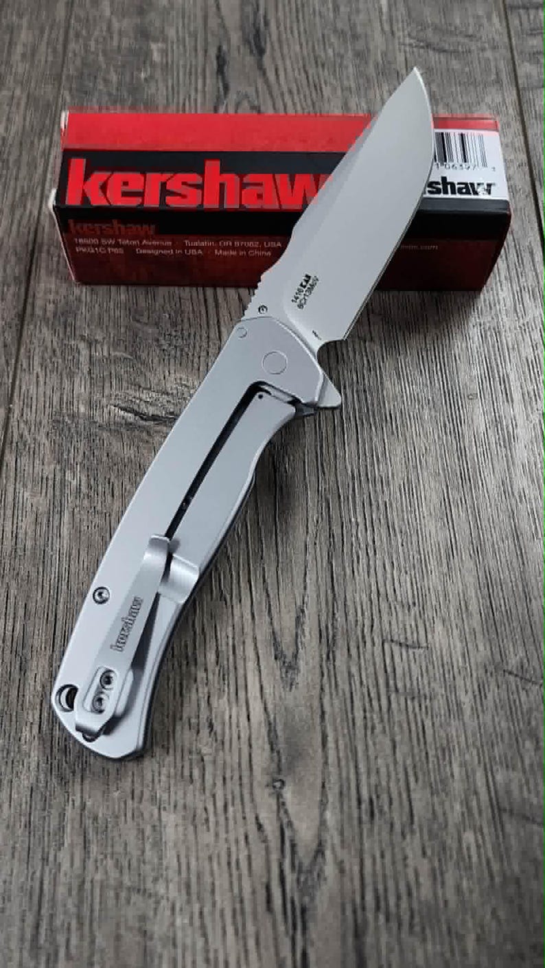May include: A silver Kershaw folding knife with a partially serrated blade, on a wooden surface. The knife has a pocket clip and the brand name is visible. A red and black box with the Kershaw logo is in the background.