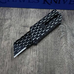 Peut inclure: Un cutter noir avec un motif de clés et une lame argentée. Le couteau est partiellement ouvert, révélant la lame et le motif de clés sur le manche. Le texte "Chaves Knife" est visible en arrière-plan.