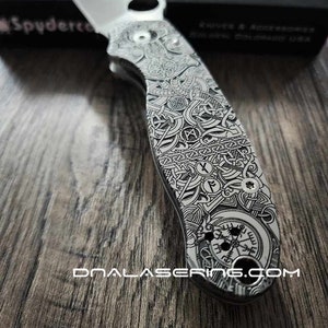 Puede incluir: Un cuchillo de bolsillo plateado con un diseño negro y blanco detallado en el mango. El diseño presenta patrones intrincados y una brújula. El cuchillo está hecho por Spyderco y está grabado con el texto "DNA Laser Engraving.com".