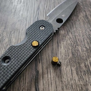 May include: A black folding knife with a partially serrated blade and a black textured handle. The knife is open and the blade is visible. There are two gold-colored screws on the handle and a gold-colored screw on the blade. A gold-colored screw is lying on the wooden surface next to the knife.
