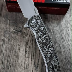 May include: A silver folding knife with an ornate, engraved floral handle. The blade is partially open. The knife is displayed on a wooden surface. The box in the background displays the brand name "Kershaw".