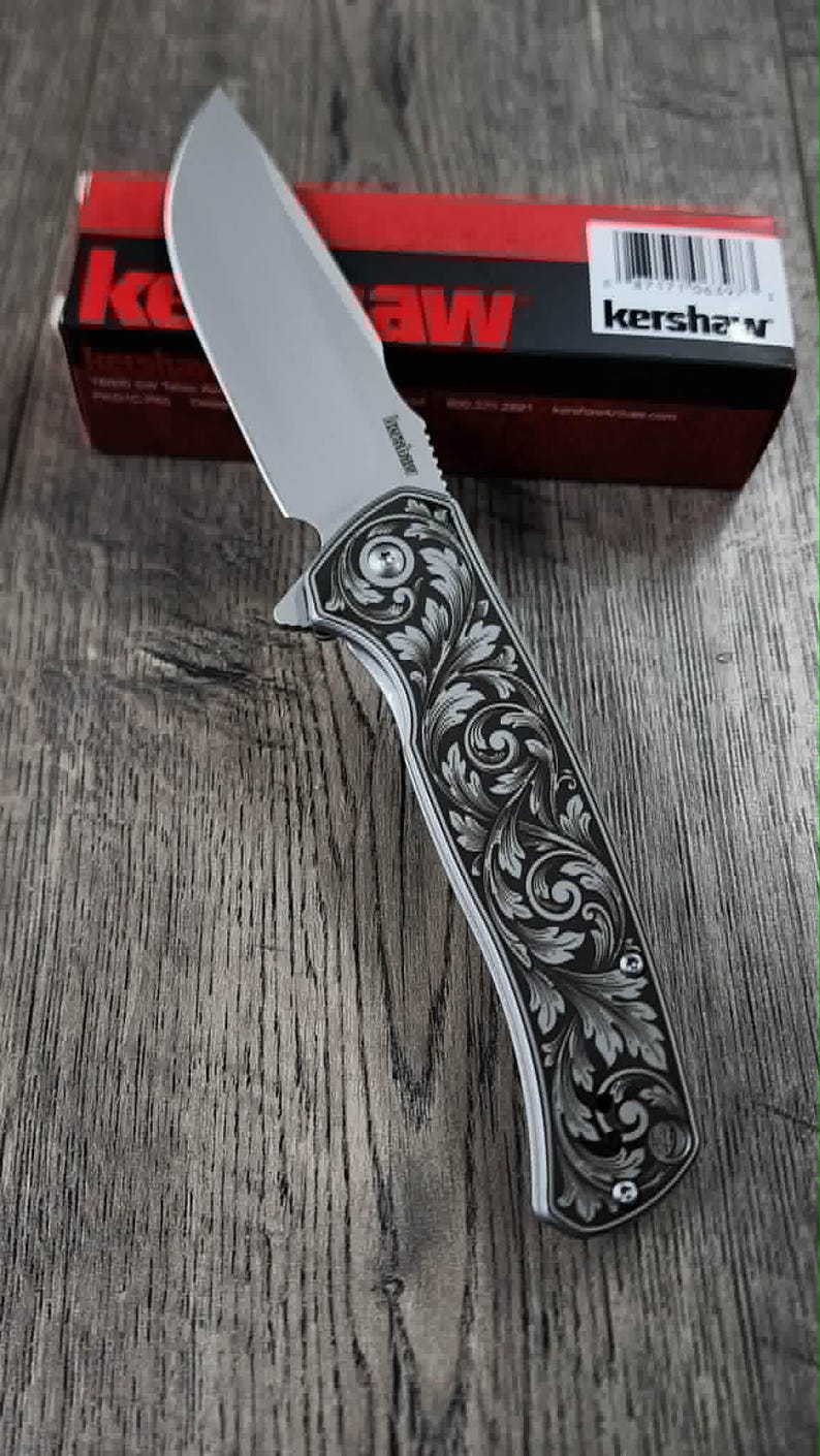 May include: A silver folding knife with an ornate, engraved floral handle. The blade is partially open. The knife is displayed on a wooden surface. The box in the background displays the brand name "Kershaw".