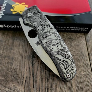 May include: A silver pocket knife with a detailed design of two women with sugar skulls. The knife has a black handle and a silver blade. The knife is closed and resting on a wooden surface.