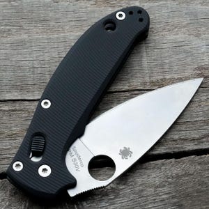 Op de afbeelding: Een zwart vouwmes met een gedeeltelijk gekarteld zilverkleurig lemmet. Het mes is open, met het lemmet met de tekst "Spyderco PM S30V" en een spinnenlogo. De handgreep is zwart met een getextureerde grip en zilveren hardware.