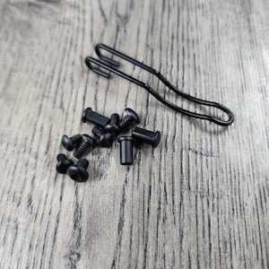 May include: A close-up of a set of black screws and a black metal hook. The screws are scattered on a light brown wooden surface.