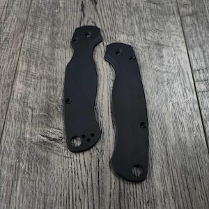 May include: Two black knife handle scales with a textured surface and screw holes. The scales are made of a durable material and are designed to fit a folding knife. The handles are shown on a wooden surface.
