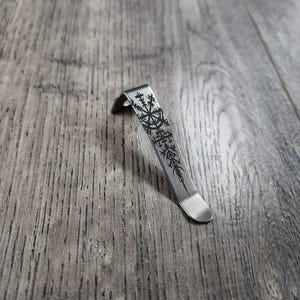 May include: A silver-colored, curved metal clip with a decorative black etched design. The clip has a rounded end and a detailed pattern, possibly a symbol or emblem. The item is shown on a wooden surface.