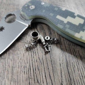 Könnte beinhalten: Ein silbernes Taschenmesser mit einem Tarnmuster-grünen Griff und dem Text "Spyderco CPM S30V" auf der Klinge. Das Messer ist geöffnet und liegt auf einer Holzoberfläche mit mehreren kleinen silbernen Schrauben, die um es herum verstreut sind.