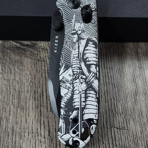 May include: A black and silver folding pocket knife with a detailed samurai warrior design. The knife features the brand name "KIZER" on the blade and a decorative handle with a black and white illustration. The knife is on a wooden surface.
