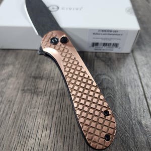 May include: A copper-coloured pocket knife with a black blade and a diamond-patterned handle. The knife is closed and has a button lock mechanism. The knife is on a wooden surface.