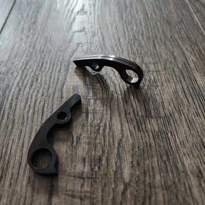 May include: Two black bottle openers are displayed on a wooden surface. Each opener has a curved design with a circular opening and a small hole. The openers are made of a dark, possibly metal material.