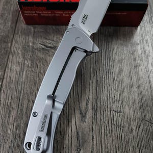 May include: A silver folding knife with a partially serrated blade, on a wooden surface. The knife has a pocket clip and the brand name "Kershaw" is visible. A red and black box with the "Kershaw" logo is in the background.