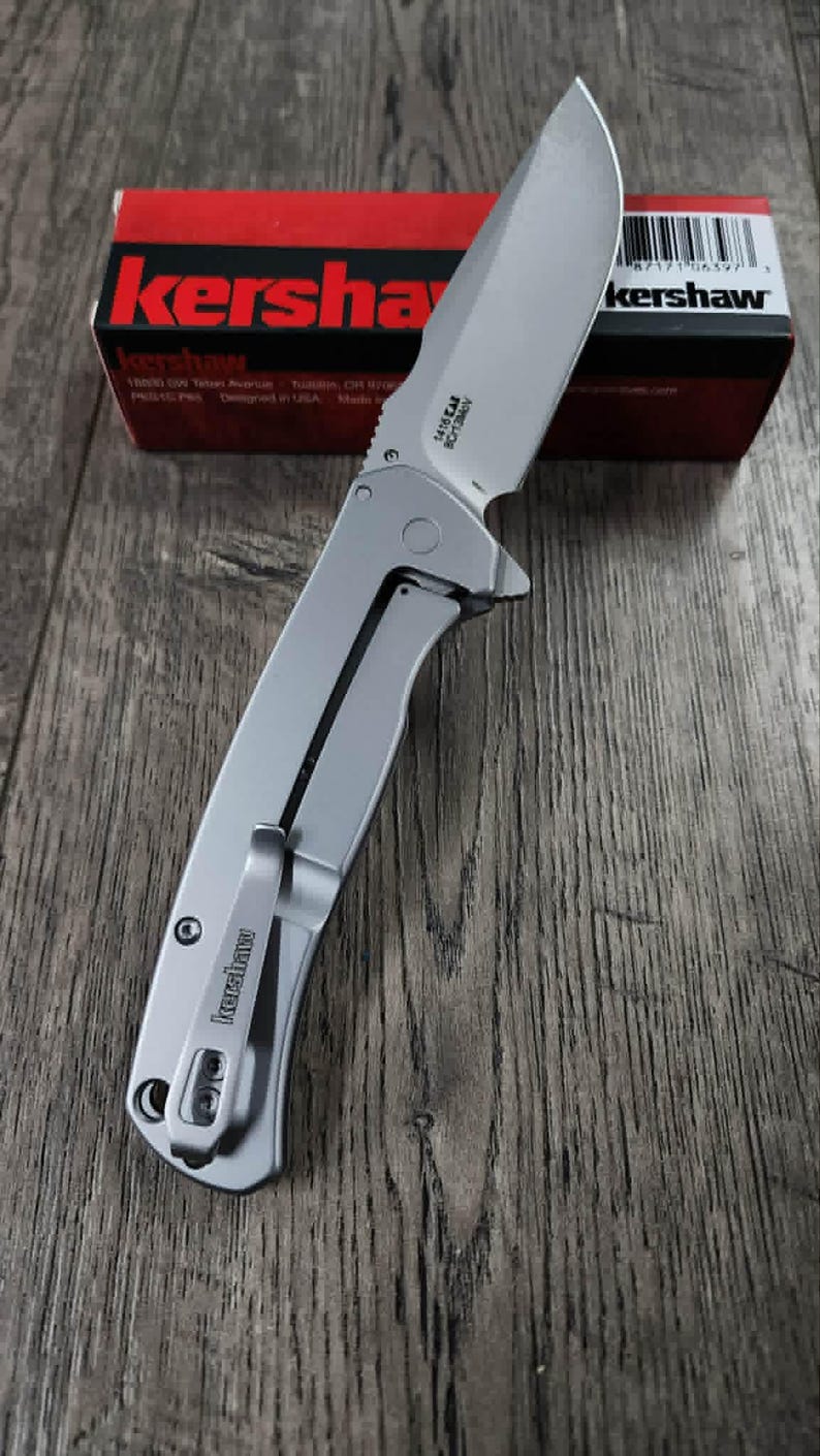 May include: A silver folding knife with a partially serrated blade, on a wooden surface. The knife has a pocket clip and the brand name "Kershaw" is visible. A red and black box with the "Kershaw" logo is in the background.