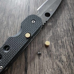 May include: A black folding knife with a partially open blade and a gold-colored accent on the handle. Two gold-colored screws and a small screw are visible on a wooden surface.