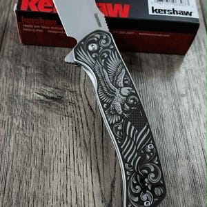 May include: A silver folding knife with a blade and a handle featuring an engraved eagle and American flag design. The knife is displayed on a wooden surface, with a red and black Kershaw box in the background.