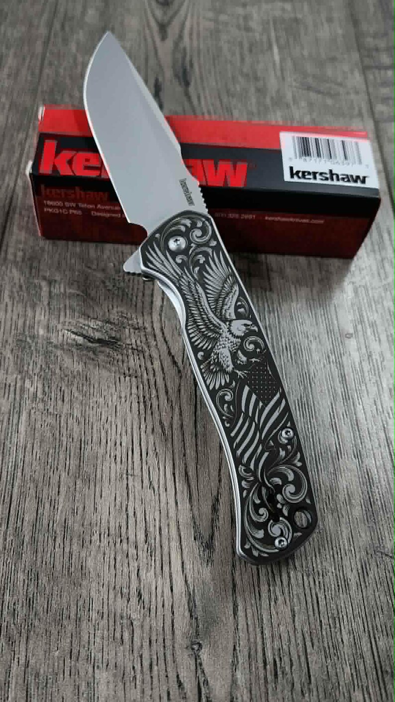 May include: A silver folding knife with a blade and a handle featuring an engraved eagle and American flag design. The knife is displayed on a wooden surface, with a red and black Kershaw box in the background.