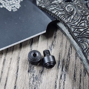 Puede incluir: Dos tornillos negros con cabeza de estrella, que descansan sobre una superficie de madera. Los tornillos se utilizan para sujetar el mango de un cuchillo a la hoja.