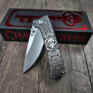 May include: A silver folding knife with detailed engraved patterns and a skull emblem. The knife is open, showing the blade. The knife is on a wooden surface, with a black box featuring the text "CHAVES KNIVES" in red.