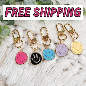May include: Five gold keychains with different colored smiley faces. The keychains are on a white fuzzy surface. The keychains are pink, black, blue, purple, and yellow.