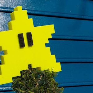 May include: A yellow star-shaped object with black eyes, made of foam blocks, is attached to a small green tree. The star is against a blue background.