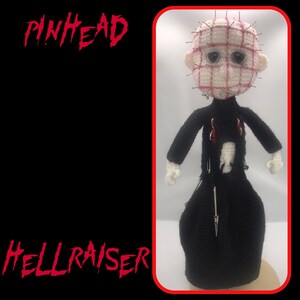 May include: A crocheted Pinhead doll from the Hellraiser series. The doll features a white head with a grid pattern and pins, black clothing, and safety pins. The words "PINHEAD" and "HELLRAISER" are written in red.