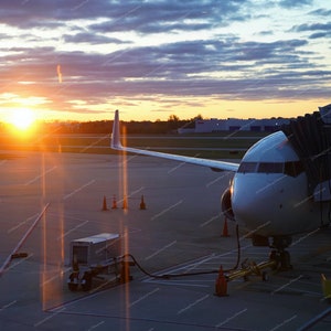 May include: A white airplane is parked at a gate at an airport. The airplane is connected to a jet bridge. The sun is setting in the background, casting a golden glow over the scene.