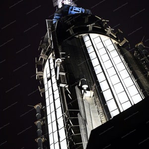 May include: A close-up view of the top of a skyscraper at night. The building is lit up with bright white lights, and the details of the structure are visible against the dark sky.