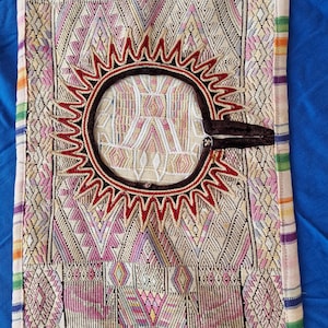May include: Textile with intricate geometric patterns in cream, pink, and purple. A central sun-like design in red and brown accents the piece. The edges feature colorful striped borders, showcasing a blend of traditional craftsmanship and vibrant colors.