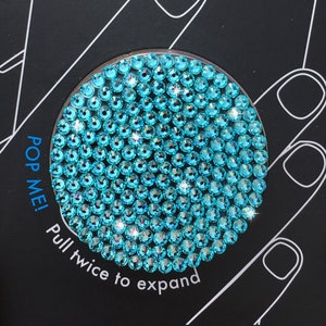 May include: A turquoise blue phone grip with a pop-out design. The grip is covered in small, sparkly rhinestones. The text "POP ME! Pull twice to expand" is printed on the black background.