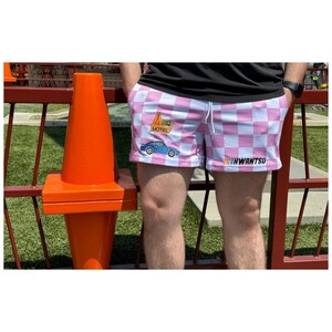 May include: Pink and white checkered shorts with a blue car and a motel sign graphic. The text on the sign says "Cozy Cone Motel". The shorts also have the text "KWANTSU" printed on the right leg.