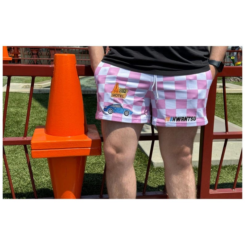 May include: Pink and white checkered shorts with a blue car and a motel sign graphic. The text on the sign says "Cozy Cone Motel". The shorts also have the text "KWANTSU" printed on the right leg.