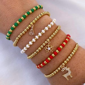 May include: A collection of gold beaded bracelets with Christmas-themed charms. The bracelets feature green, white, and red beads. Charms include a Christmas tree, snowflake, and a reindeer. The bracelets are worn on a wrist.