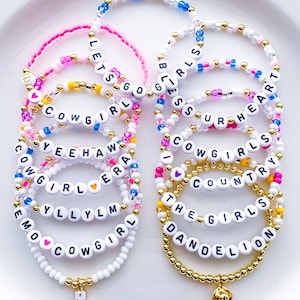 May include: A collection of beaded bracelets in various colors, including pink, white, gold, and blue. Some bracelets feature letter beads spelling out phrases like "Cowgirl" and "Let's Go." One bracelet has a boot charm, and another has a cowboy hat charm.