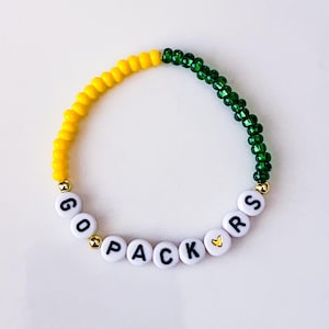 May include: A beaded bracelet with yellow and green beads. The bracelet spells out "GO PACKERS" with white letter beads. Gold-colored beads separate the words and the colors.