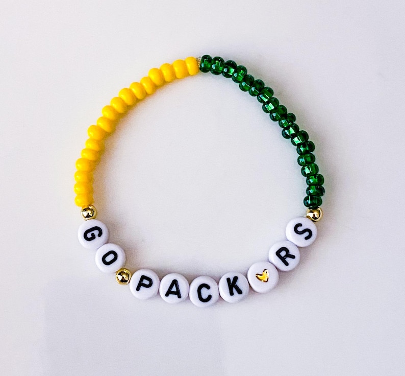 May include: A beaded bracelet with yellow and green beads. The bracelet spells out "GO PACKERS" with white letter beads. Gold-colored beads separate the words and the colors.