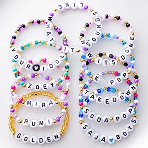 Pode incluir: Uma cole&ccedil;&atilde;o de pulseiras de contas com contas de letras brancas que soletram palavras e nomes. As pulseiras apresentam v&aacute;rias cores de contas, incluindo ouro, rosa, roxo e azul. Algumas pulseiras t&ecirc;m pingentes de cora&ccedil;&atilde;o e estrela.