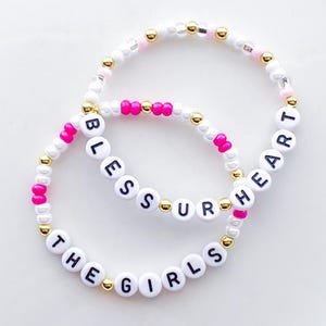 Puede incluir: Dos pulseras de cuentas con cuentas blancas, rosas y doradas. Una pulsera deletrea "BLESS UR HEART" y la otra dice "THE GIRLS" en letras negras sobre cuentas blancas. Un accesorio elegante.